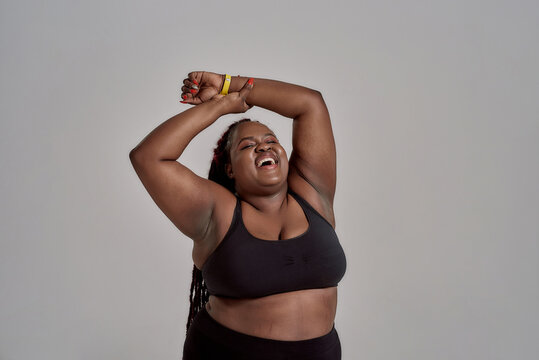 Love my body. Plump, plus size african american woman in sportswear laughing, lifting her arms up in studio over grey background. Concept of sport, healthy lifestyle, body positive, equality