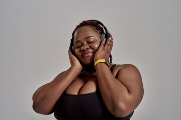 Music. Plump, plus size african american woman in sportswear and headphones listening to music, standing in studio over grey background. Concept of sport, healthy lifestyle, body positive, equality