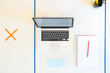 Top view of a desk with a laptop, notebook, medical face mask, and social distancing measures to prevent the spread of the coronavirus.