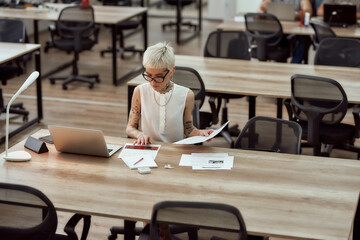 Young and cute tattoed business woman with short haircut analyzing documents and working on laptop...