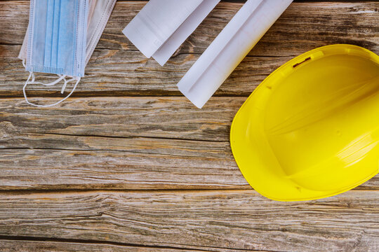 The Coronavirus prevention face mask protection is on builder of architects blueprints working on engineering office construction plans on yellow safety helmet hard hat