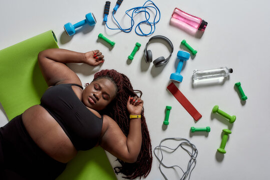 Better change. Top view of tired plump, plus size african american woman in sportswear lying on yoga mat after workout with sports, fitness equipment and headphones near her over grey background - Powered by Adobe