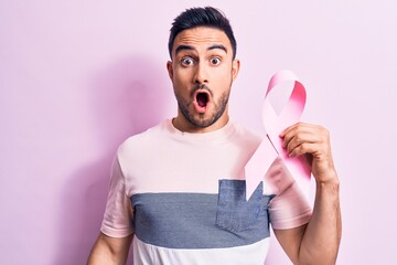 Young handsome man with beard holding pink cancer ribbon symbol over isolated background scared and amazed with open mouth for surprise, disbelief face