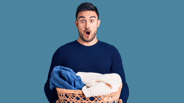 Young handsome man holding laundry basket scared and amazed with open mouth for surprise, disbelief face
