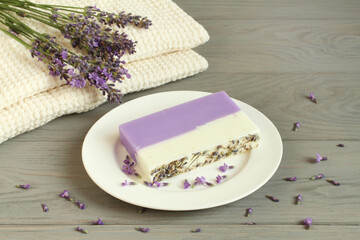 Lavender flowers on linen towels and soap made of lavender, olive oils and cocoa butter on a small saucer are located on a gray wooden table near a white brick wall. Closeup.