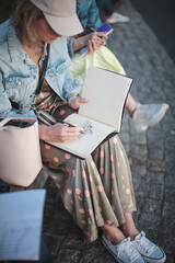 Obraz premium Young beautiful girl draws in a park in a group. Outline in a sketchbook.