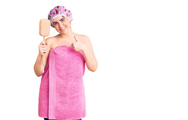 Young caucasian woman wearing shower cap and towel holding sponge smiling happy and positive, thumb up doing excellent and approval sign