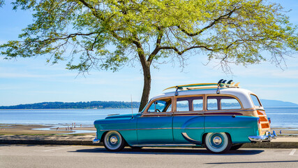 Old classic retro car with surfboards and beach tool on the roof of the car, at shade underneath a beautiful green-leafed tree, with people on the beach in the background. A commonplace, California.