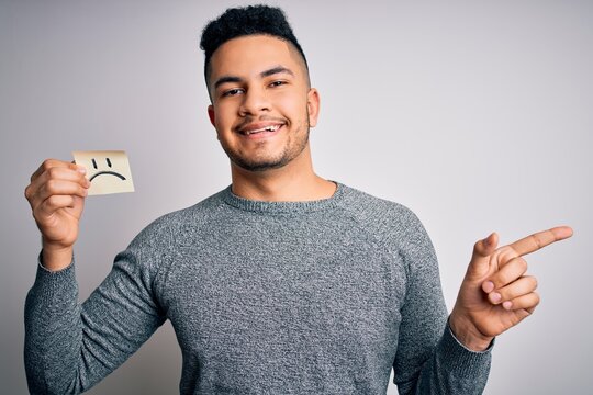 Handsome man holding reminder paper with sad emotion face emoji over white background very happy pointing with hand and finger to the side