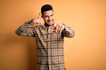 Young handsome man wearing casual shirt standing over isolated yellow background smiling doing talking on the telephone gesture and pointing to you. Call me.