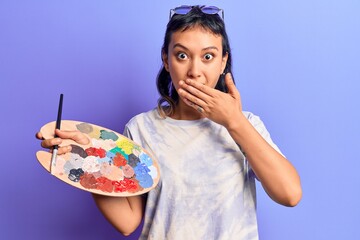 Young woman holding paintbrush and palette covering mouth with hand, shocked and afraid for...