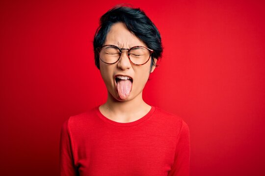 Young beautiful asian girl wearing casual t-shirt and glasses over isolated red background sticking tongue out happy with funny expression. Emotion concept.