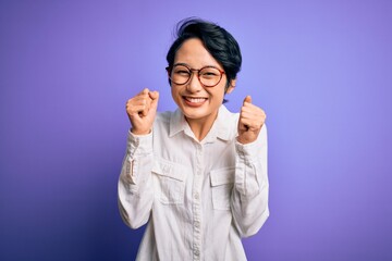 Young beautiful asian girl wearing casual shirt and glasses standing over purple background excited for success with arms raised and eyes closed celebrating victory smiling. Winner concept.