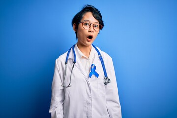 Fototapeta premium Young beautiful asian doctor girl wearing stethoscope and coat with blue cancer ribbon afraid and shocked with surprise expression, fear and excited face.