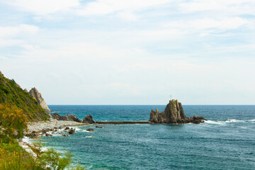 Coastal view of a rocky beach in Moneglia, Liguria, Italy.  narrow shoreline a is connected to a giant sea rock islet with a man made concrete path.  The islet harbors stairs and a giant cross on top.
