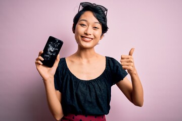 Young beautiful chinese woman holding broken smartphone showing craked screen happy with big smile...
