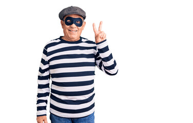 Senior handsome man wearing burglar mask and t-shirt smiling with happy face winking at the camera doing victory sign with fingers. number two.