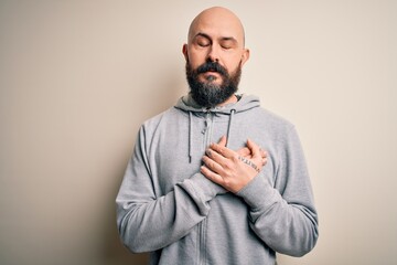 Handsome sporty bald man with beard wearing sweatshirt standing over pink background smiling with hands on chest with closed eyes and grateful gesture on face. Health concept.