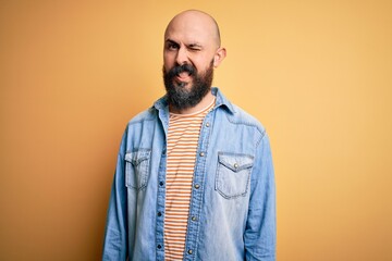Handsome bald man with beard wearing casual denim jacket and striped t-shirt winking looking at the...