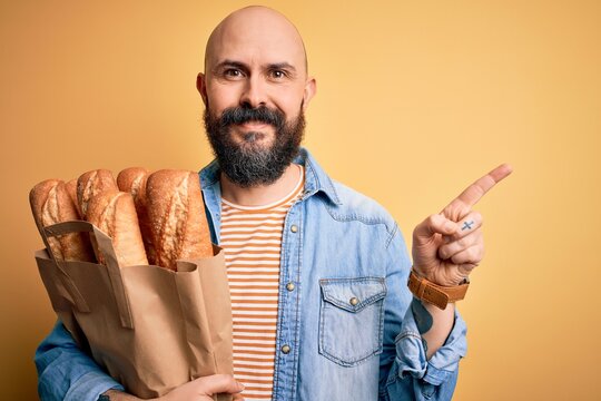 Handsome bald man with beard holding paper bag with bread over yellow background very happy pointing with hand and finger to the side