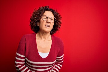 Middle age beautiful curly hair woman wearing casual striped sweater over red background with hand on stomach because nausea, painful disease feeling unwell. Ache concept.