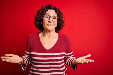 Middle age beautiful curly hair woman wearing casual striped sweater over red background clueless and confused expression with arms and hands raised. Doubt concept.