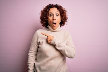 Middle age beautiful curly hair woman wearing casual turtleneck sweater over pink background Surprised pointing with finger to the side, open mouth amazed expression.