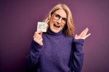 Middle age beautiful blonde woman wearing glasses holding reminder paper with yes message very...