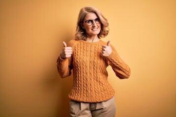 Middle age beautiful blonde woman wearing casual sweater and glasses over yellow background success sign doing positive gesture with hand, thumbs up smiling and happy. Cheerful expression and winner