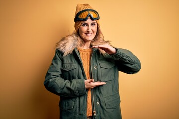 Middle age beautiful blonde skier woman wearing snow sportwear and ski goggles gesturing with hands showing big and large size sign, measure symbol. Smiling looking at the camera. Measuring concept.