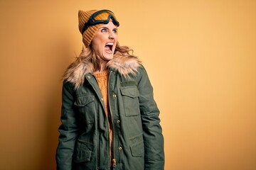 Middle age beautiful blonde skier woman wearing snow sportwear and ski goggles angry and mad screaming frustrated and furious, shouting with anger. Rage and aggressive concept.