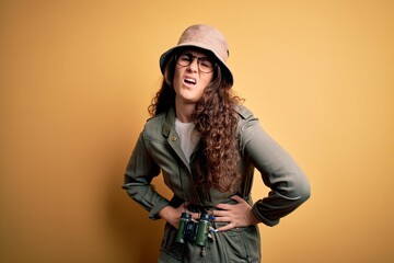 Young beautiful tourist woman on vacation wearing explorer hat and binoculars with hand on stomach...