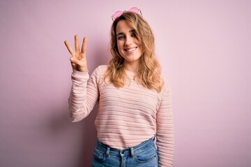 Young beautiful blonde woman wearing casual sweater and sunglasses over pink background showing and pointing up with fingers number three while smiling confident and happy.