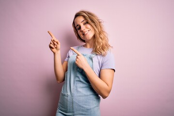 Young beautiful blonde woman wearing casual denim overalls standing over pink background smiling and looking at the camera pointing with two hands and fingers to the side.