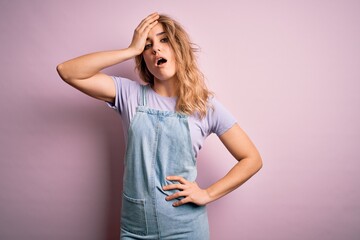 Young beautiful blonde woman wearing casual denim overalls standing over pink background surprised with hand on head for mistake, remember error. Forgot, bad memory concept.