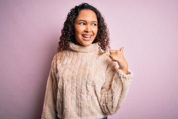 Young beautiful woman with curly hair wearing casual sweater standing over pink background smiling with happy face looking and pointing to the side with thumb up.