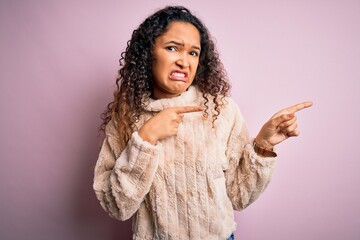 Young beautiful woman with curly hair wearing casual sweater standing over pink background Pointing aside worried and nervous with both hands, concerned and surprised expression