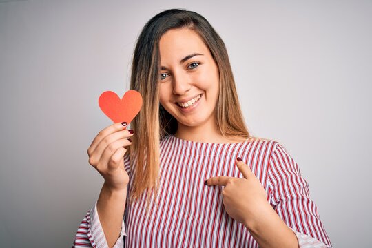 Young beautiful blonde romantic woman holding red paper heart over white background with surprise face pointing finger to himself