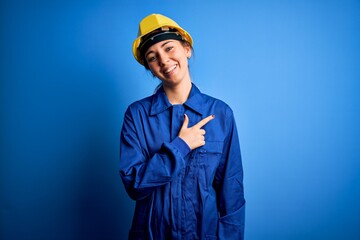 Young beautiful worker woman with blue eyes wearing security helmet and uniform cheerful with a smile of face pointing with hand and finger up to the side with happy and natural expression on face