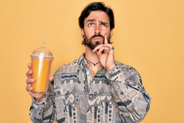Young handsome hispanic bohemian hipster man with bear drinking healthy fresh orange juice serious...