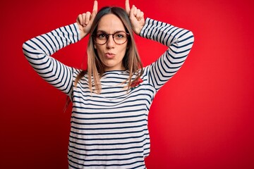 Young beautiful blonde woman with blue eyes wearing glasses standing over red background doing funny gesture with finger over head as bull horns