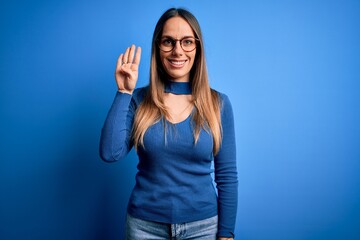 Obraz na płótnie Canvas Young beautiful blonde woman with blue eyes wearing glasses standing over blue background showing and pointing up with fingers number four while smiling confident and happy.