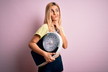 Young beautiful blonde sporty woman on diet holding weight machine over pink background serious face thinking about question, very confused idea