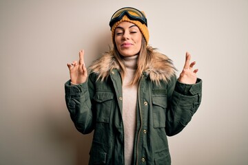 Young brunette skier woman wearing snow clothes and ski goggles over white background gesturing finger crossed smiling with hope and eyes closed. Luck and superstitious concept.
