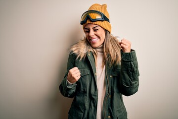 Young brunette skier woman wearing snow clothes and ski goggles over white background celebrating surprised and amazed for success with arms raised and eyes closed. Winner concept.
