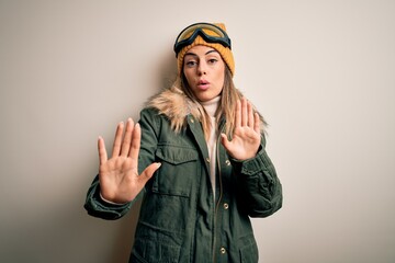 Young brunette skier woman wearing snow clothes and ski goggles over white background Moving away hands palms showing refusal and denial with afraid and disgusting expression. Stop and forbidden.