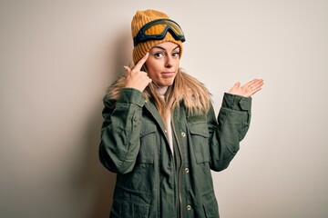 Young brunette skier woman wearing snow clothes and ski goggles over white background confused and annoyed with open palm showing copy space and pointing finger to forehead. Think about it.