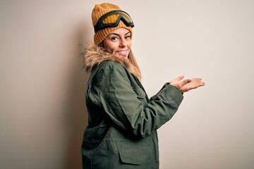 Young brunette skier woman wearing snow clothes and ski goggles over white background pointing aside with hands open palms showing copy space, presenting advertisement smiling excited happy