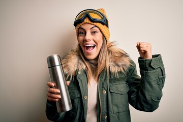 Young beautiful brunette skier woman wearing ski goggles drinking thermo with coffe screaming proud...