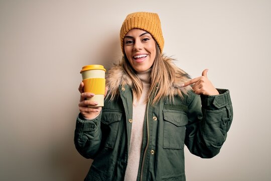 Young beautiful brunette woman wearing snow clothes drinking cup of coffee with surprise face pointing finger to himself